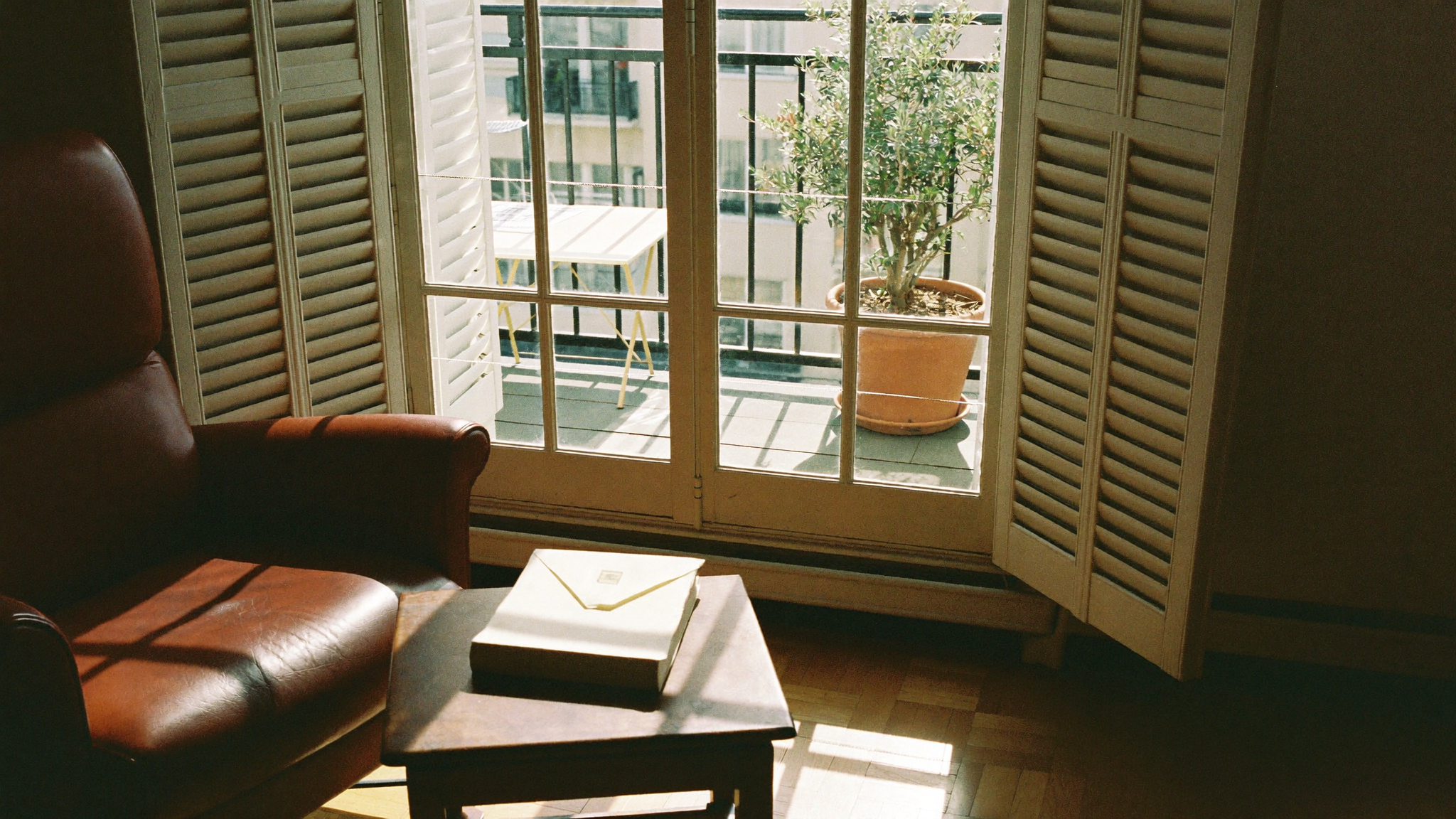 The diaspora property pattern — an empty Greek apartment in Glyfada with shuttered balcony, leather armchair, and an unopened envelope on the side table, awaiting an owner who lives abroad